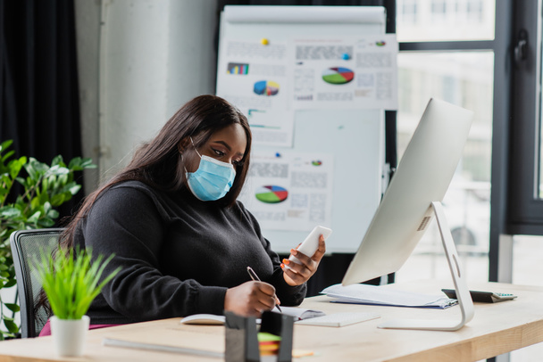 african american plus size businesswoman in medical mask holding smartphone and writing in notebook  - 写真・画像