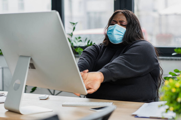 cansado africano americano más tamaño mujer de negocios en médico máscara estiramiento cerca de ordenador monitor  - Foto, imagen