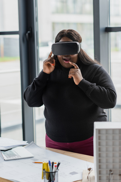 κατάπληκτος Αφροαμερικανός συν μέγεθος γυναίκα σε VR ακουστικά κοντά στο μοντέλο χάρτινο σπίτι και το σχέδιο στο γραφείο - Φωτογραφία, εικόνα