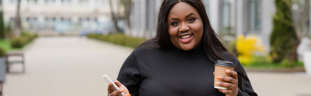 happy african american plus size woman holding coffee to go and smartphone outside, banner - Zdjęcie, obraz