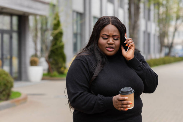 Αφροαμερικάνα συν μέγεθος γυναίκα κρατώντας καφέ για να πάει και μιλώντας στο smartphone έξω  - Φωτογραφία, εικόνα