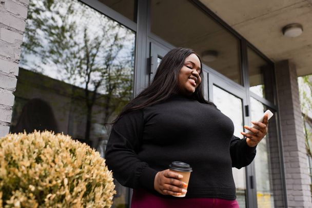 low angle view of cheerful african american plus size woman holding coffee to go and looking at smartphone outside  - Foto, afbeelding