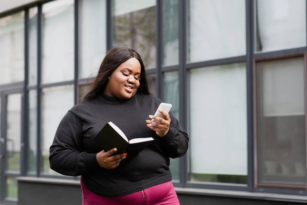χαρούμενα Αφρικής Αμερικής συν μέγεθος γυναίκα κρατώντας σημειωματάριο και smartphone έξω  - Φωτογραφία, εικόνα