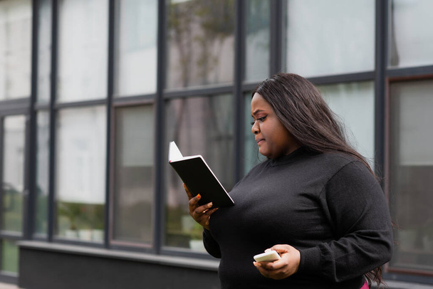 Αφροαμερικάνα συν μέγεθος γυναίκα κρατώντας σημειωματάριο και smartphone έξω  - Φωτογραφία, εικόνα