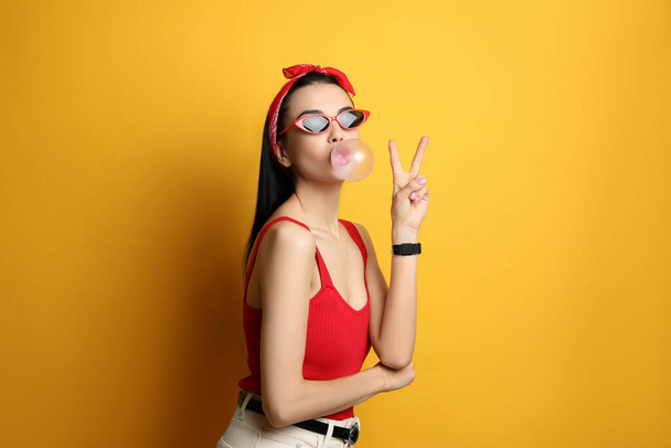 Fashionable young woman in pin up outfit blowing bubblegum on yellow background - Foto, Bild