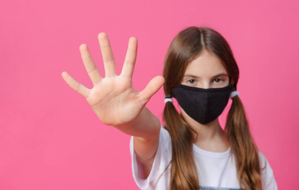 white teenage girl in a black mask shows a stop sign with her hand on a pink background - Фото, изображение