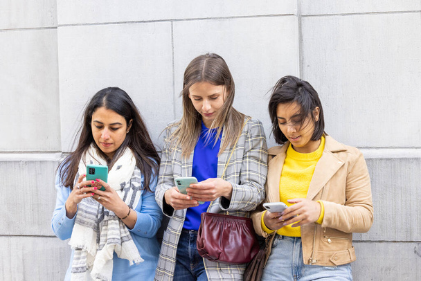 Grupo multiétnico de hermosas amigas jóvenes de pie contra una pared usando teléfonos inteligentes móviles - Foto, Imagen