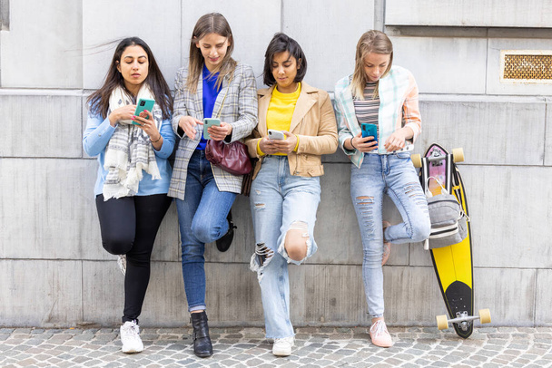 Grupo multiétnico de hermosas amigas jóvenes de pie contra una pared usando teléfonos inteligentes móviles - Foto, imagen