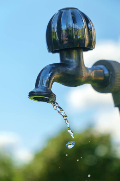 Pouring fresh water  from the metal tap with blurred nature and blue cloudy sky background, selective focus - Foto, imagen