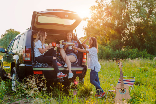Enjoying the rest and socializing on a picnic trip with your best friends. A group of young cheerful women travel in nature by car. Hipster girls laugh at sunset. celebrate - Photo, image