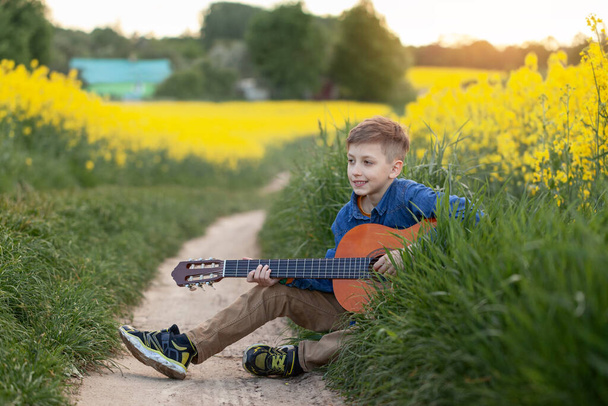 Portrait of cute boy playing a guitar sitting on road in the summer yellow field. - 写真・画像