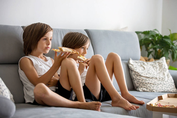 Niños lindos, sentados en el sofá, comiendo pizza y viendo televisión. Niño hambriento tomando un bocado de pizza en un día de fiesta de pizza en casa - Foto, Imagen