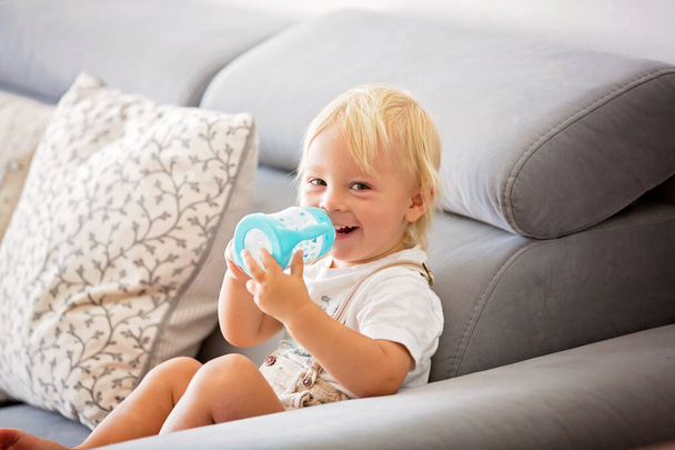 Adorable baby boy drinking milk from a bottle in a white sunny living room. Formula drink for infant. - Photo, Image