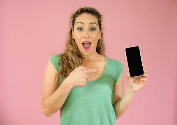 Excited young woman pointing mobile phone isolated over pink background. - Foto, Imagem
