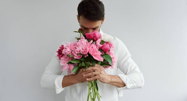Вид спереди на красивого мужчину с букетом розовых пионов в качестве подарка на День Святого Валентина или свадебного торжества. Мужчина в белой рубашке, обнюхивающий цветы в руках, изолированный на сером фоне. - Фото, изображение