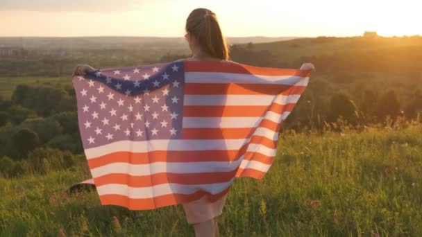Πίσω όψη της ευτυχισμένης γυναίκας με την εθνική σημαία των ΗΠΑ να κάθεται σε εξωτερικούς χώρους το ηλιοβασίλεμα. Θετικό κορίτσι γιορτάζει την ημέρα ανεξαρτησίας των Ηνωμένων Πολιτειών. Διεθνής ημέρα της έννοιας της δημοκρατίας. - Πλάνα, βίντεο