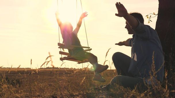 Маленькая девочка качается на качелях на закате и машет руками в полете, парить, как самолет над землей, замедленная съемка, счастливая семья, командная работа, играть с ребенком на открытом воздухе вместе - Фото, изображение