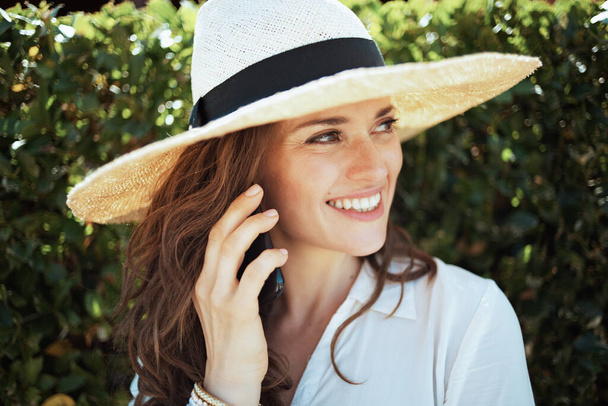 happy elegant housewife in white shirt with hat using a smartphone outdoors near green wall. - 写真・画像
