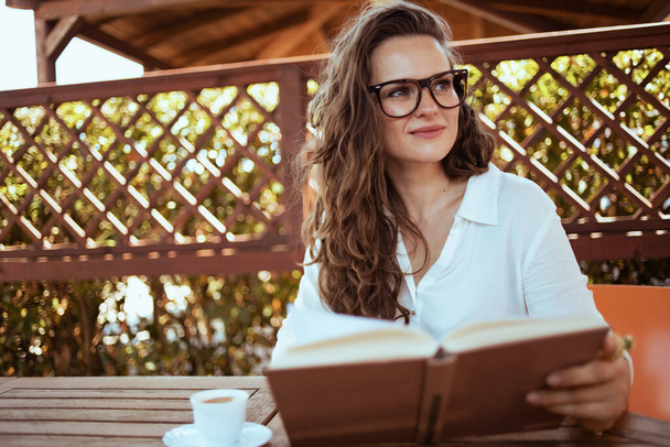 pensive elegant 40 years old woman in white shirt with cup of coffee, book and eyeglasses sitting at the table in the terrace. - Photo, image