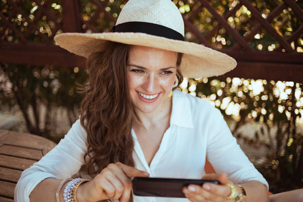 smiling trendy 40 years old woman in white shirt with hat sending text message using smartphone in the patio of guest house hotel. - Фото, изображение