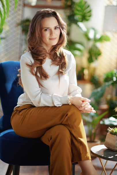 Green Home. Portrait of elegant 40 years old housewife with long wavy hair in the modern living room in sunny day in green pants and grey blouse sitting in a blue armchair. - Фото, изображение