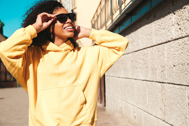 Beautiful black woman with afro curls hairstyle.Smiling hipster model in yellow hoodie. Sexy carefree female posing on the street background in sunglasses. Cheerful and happy - Foto, Imagen