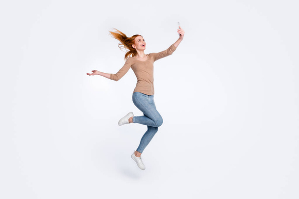 Full length photo of sweet funky redhair woman beige shirt jumping high tacking selfie modern device isolated white color background - 写真・画像