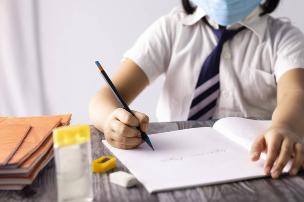 Focus selettivo a portata di mano di un bambino indiano che scrive con la matita sul taccuino indossando uniforme scolastica e maschera nasale - Foto, immagini