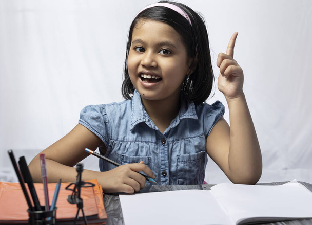 Een mooi Indiaas meisje kind denken en kijken naar de camera tijdens het studeren met een glimlachend gezicht - Foto, afbeelding