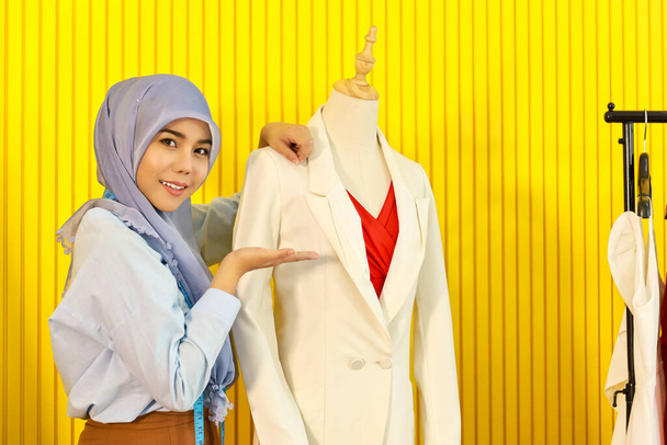 Πορτρέτο του όμορφου χαμογελαστού Ασιάτη σχεδιάστρια μουσουλμάνων με χιτζάμπ παρουσιάζει το μπουφάν και το φόρεμα της και ψάχνει για κάμερα στο σπίτι της στο στούντιο με μέτρηση ταινίας. - Φωτογραφία, εικόνα