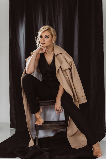 Fashion portrait of a stylish blonde in an elegant beige trench coat and black suit with smoky eye makeup on a black background. Soft selective focus. - Фото, зображення