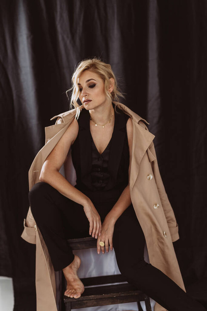 Fashion portrait of a stylish blonde in an elegant beige trench coat and black suit with smoky eye makeup on a black background. Soft selective focus. - Foto, Imagem