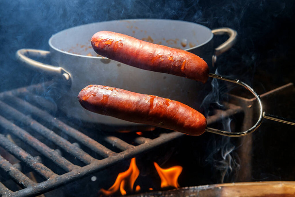 Asar bratwurst fogata en tenedor de perrito caliente con frijoles vaqueros slow cooking en segundo plano - Foto, Imagen