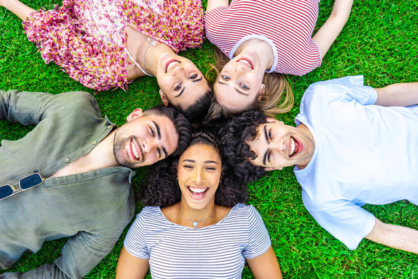 Grupo de cinco felizes jovens multirraciais gen z amigos deitado em círculo cabeça a cabeça na grama de um gramado parque olhando para a câmera a partir do fundo. Conceito de amizade sem preconceitos - Foto, Imagem