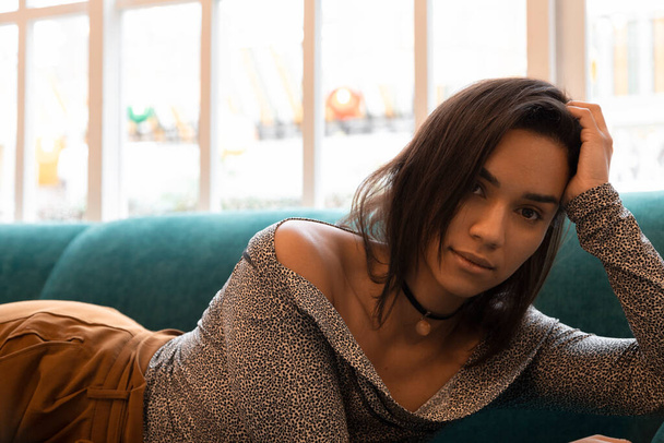 attraktive und sinnliche brünette Frau, junge lateinamerikanische Frau mit kurzen Haaren, die tagsüber auf einem Sofa liegt, Lifestyle und Mode im Studio - Foto, Bild