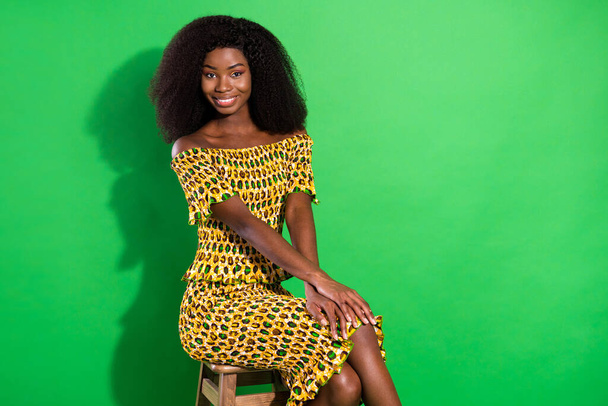 Photo of gorgeous satisfied dark skin lady sit hands on knee beaming smile isolated on green color background - 写真・画像
