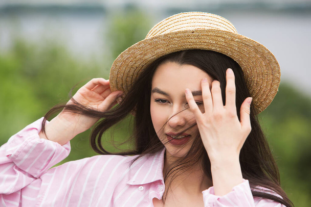Дівчина в солом'яному капелюсі вирівнює волосся рукою. Вітряна погода в сонячний літній день
. - Фото, зображення