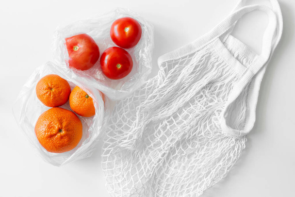 White mesh bag, vegetables, fruits flat lay on white background top view. Eco friendly, reusable shopping bag. Oranges, tomatoes in plastic bags and cotton knitted string bag. Zero waste concept. - Photo, Image