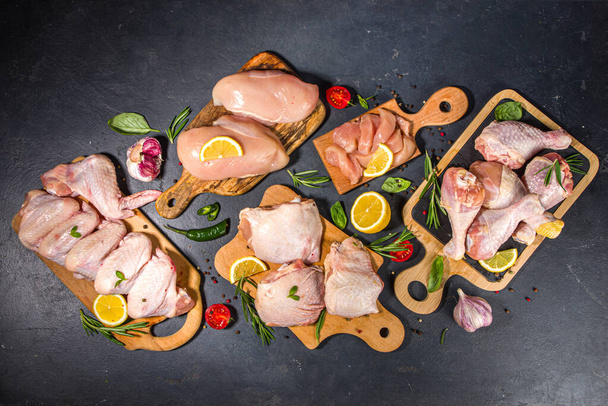 Различные порции сырого куриного мяса. Набор сырой филе курицы, бедро, крылья, полоски и ноги на черном фоне стола приготовления пищи со специями  - Фото, изображение