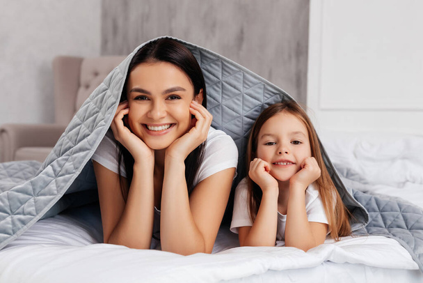 Joyful young woman resting with her pleasant child while being covered with soft blanket - Photo, image