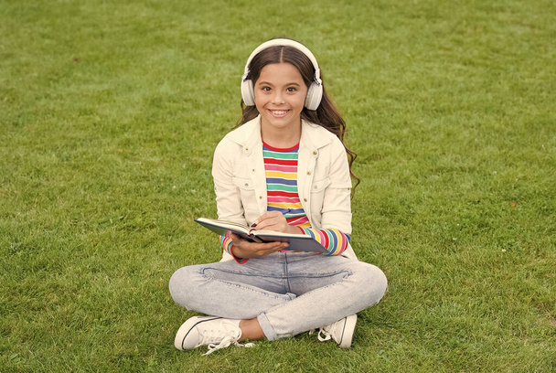 Idea concept. Child listen music on green grass. Healthy lifestyle and relax. girl write memory in diary. small kid in earphones. summer outdoor activity. enjoy spring nature. child study audio book - Fotoğraf, Görsel