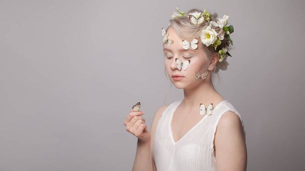 Pretty female model woman with spring flowers in her hairstyle and white butterfly sitting on her hand on white - Fotó, kép