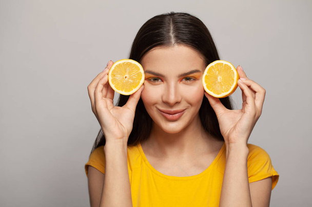 Happy brunette woman with clear skin and straight hair wearing yellow t-shirt holding lemon fruits on white background. Diet, skincare and facial treatment concept - Photo, Image