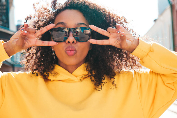 Beautiful black woman with afro curls hairstyle.Smiling hipster model in yellow hoodie. Sexy carefree female posing on the street background in sunglasses. Cheerful and happy - Foto, afbeelding