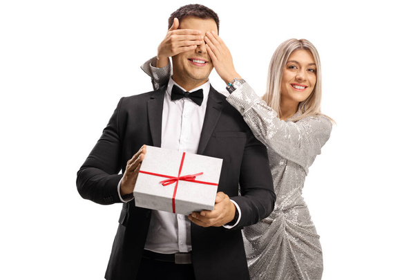 Young woman in elegant dress surprising a man with a present and covering his eyes isolated on white background - Valokuva, kuva