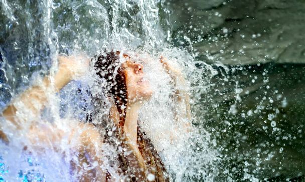 schöne junge sexy rothaarige Frau unter dem plätschernden Wasser des Wasserfalls im Spa Wellness-Pool und hebt die Arme, Spaß haben - Foto, Bild