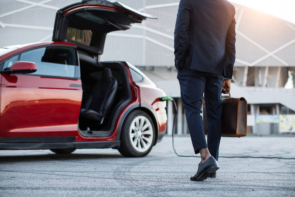 Man with suitcase standing near electric car that charging - Фото, изображение