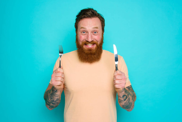 Glücklicher Mann mit Tätowierungen steht mit Besteck in der Hand zum Essen bereit - Foto, Bild