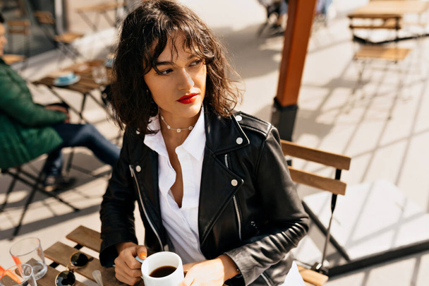 Stylish spectacular girl with red lips wearing leather jacket sitting on the summer terrace in warm sunny day. High quality photo - Photo, Image