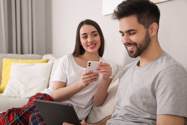 Happy couple in pajamas with gadgets on sofa at home - Foto, imagen